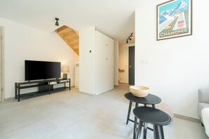 A modern living room with a flat-screen TV on a black stand, a wooden bowl on a small table, and a staircase leading to another room. - Cimes - Cozy Apartment | 5 guests | Family-friendly | Quiet (Chamonix)