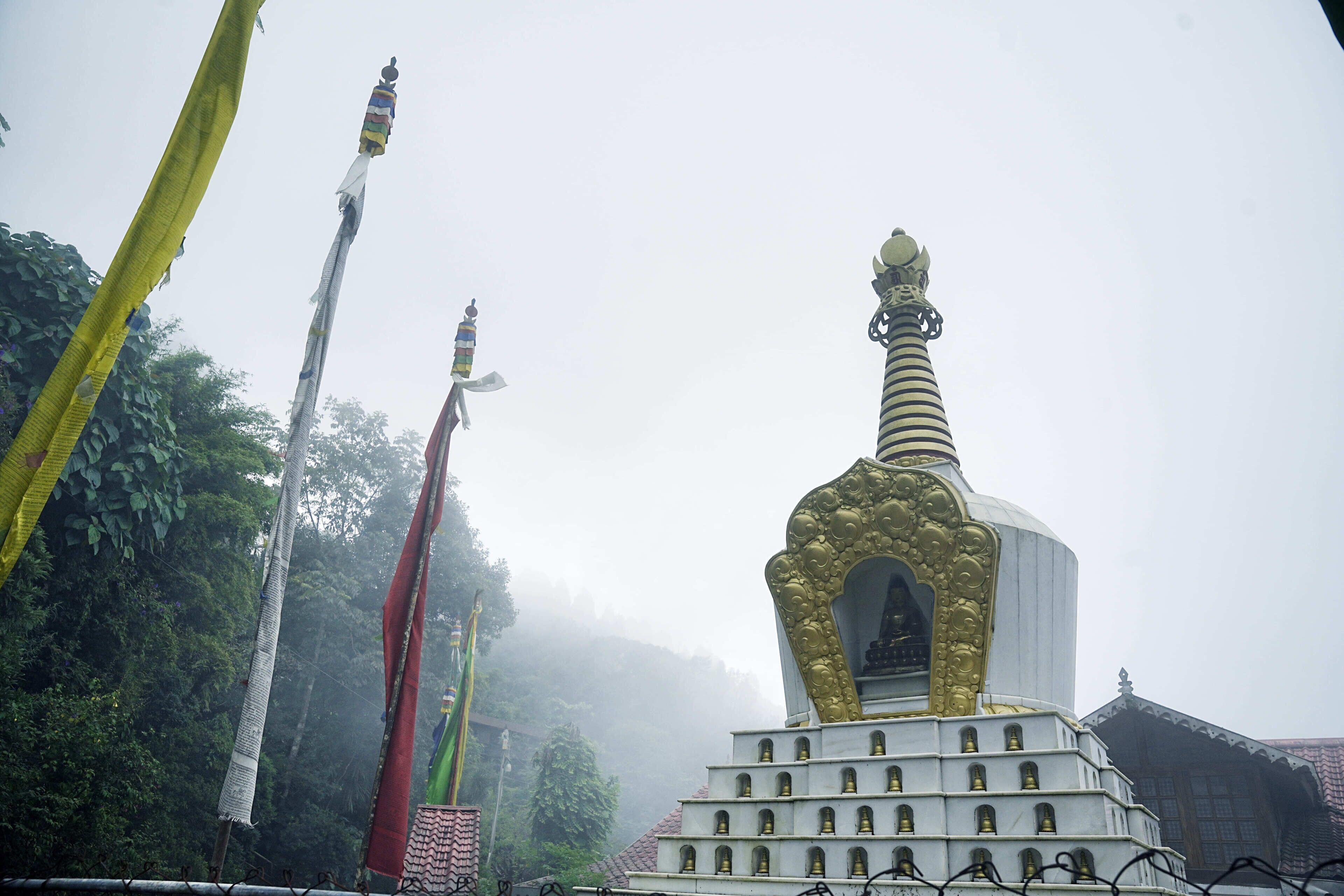 Eine große, weiße und goldene Stupa mit gestufter Basis und einer Spitze, umgeben von Bäumen und Nebel.