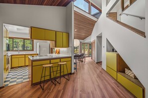 A modern kitchen with yellow cabinets, a central island with stools, and a dining area with a view of greenery. - Mid Century Modern with Sweeping Golf View (Sunriver)