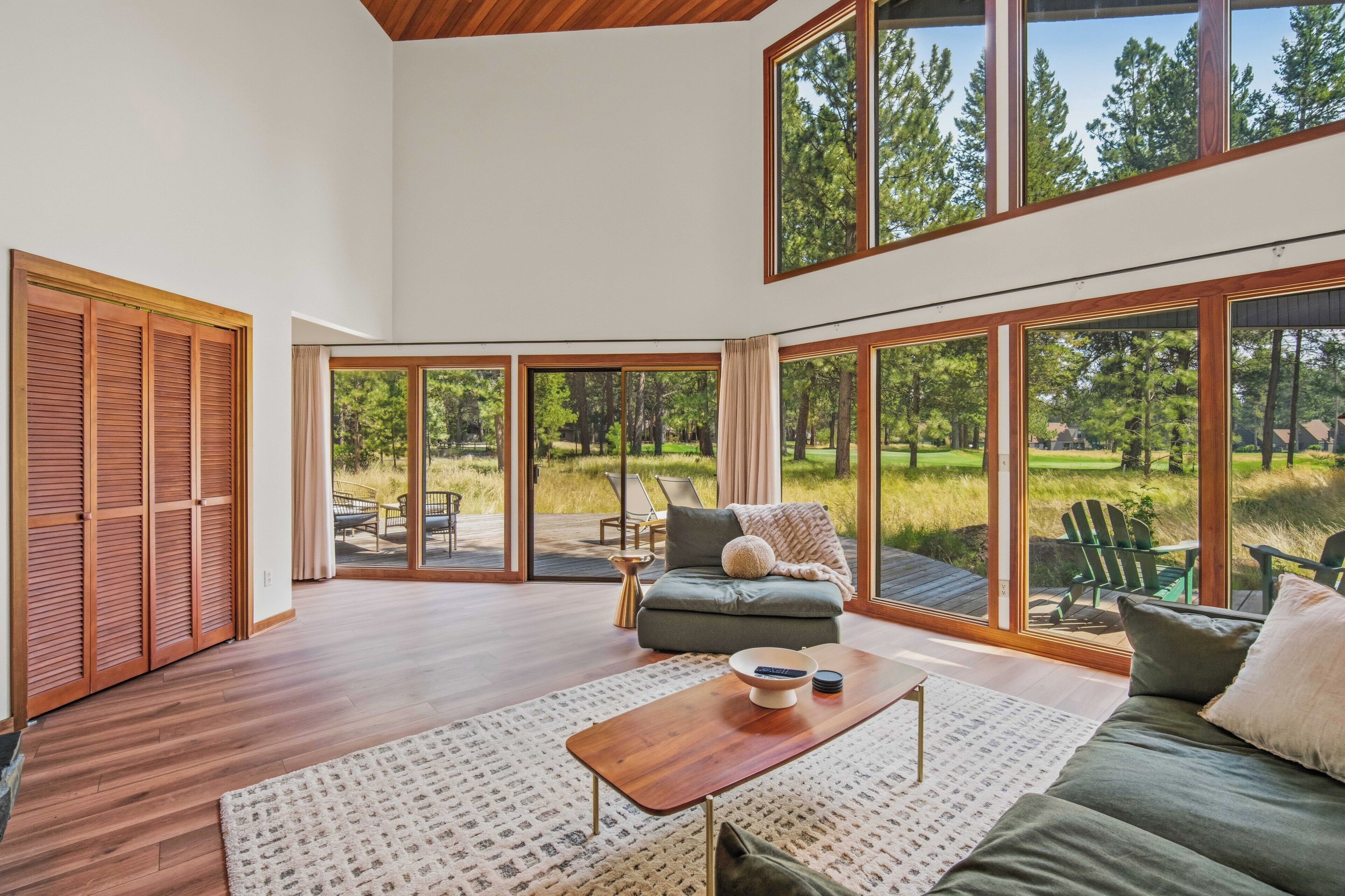 A spacious living room with a large sliding glass door leading to an outdoor deck, a sofa, a coffee table, and a view of a forested area.