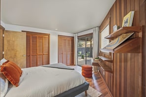 A bedroom with a bed, wooden doors, a sliding glass door to a balcony, and a shelf with books. - Mid Century Modern with Sweeping Golf View (Sunriver)
