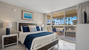 A hotel room with a bed, a nightstand, a lamp, a painting, and a view of the outside. - South Maui Beach Getaway | 2BR Condo Across from Kamaole Beach II (Kihei)