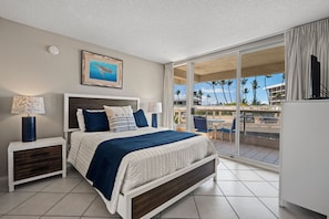 A hotel room with a bed, a nightstand, a lamp, a painting, and a view of the outside. - South Maui Beach Getaway | 2BR Condo Across from Kamaole Beach II (Kihei)