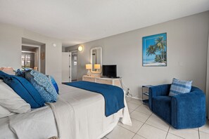 A hotel room with a bed, a desk with a TV, a chair, and a painting of a beach scene. - South Maui Beach Getaway | 2BR Condo Across from Kamaole Beach II (Kihei)