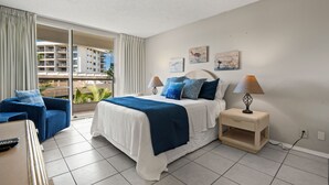 A hotel room with a bed, a nightstand, a lamp, a chair, and a view of a building through a window. - South Maui Beach Getaway | 2BR Condo Across from Kamaole Beach II (Kihei)