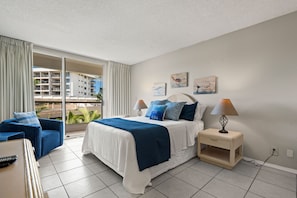 A hotel room with a bed, a nightstand, a lamp, a chair, and a view of a building through a window. - South Maui Beach Getaway | 2BR Condo Across from Kamaole Beach II (Kihei)