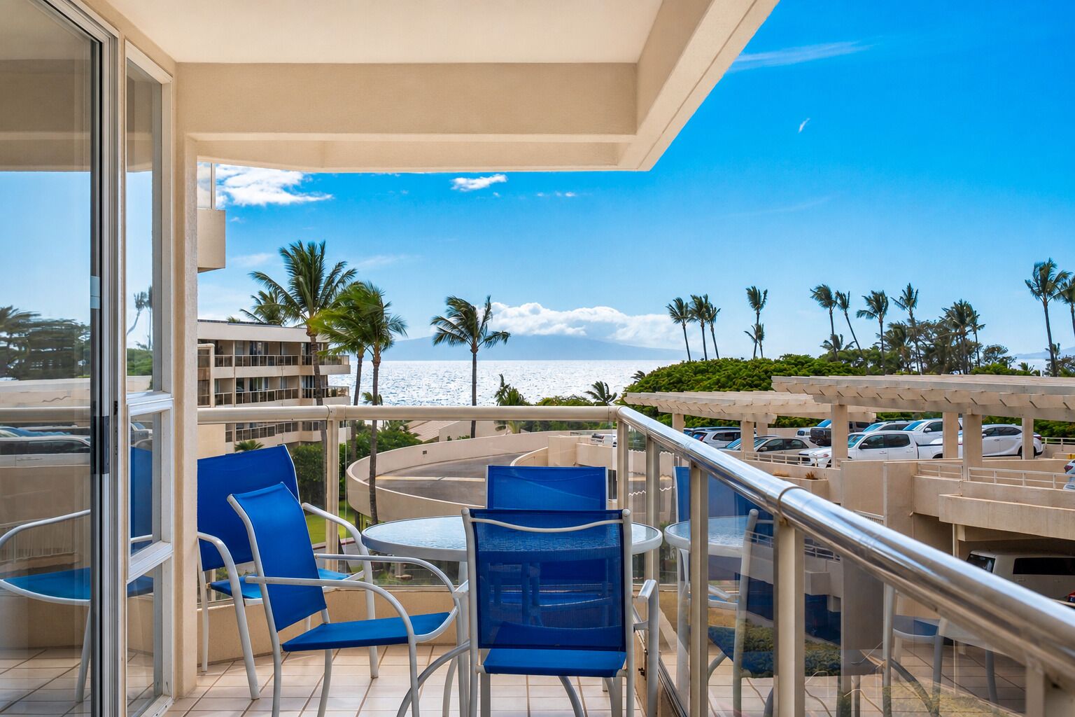 A balcony with a view of the ocean, palm trees, and a parking area.