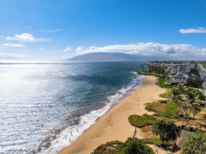 A coastal view with a beach, ocean, and a mountain in the distance. - South Maui Beach Getaway | 2BR Condo Across from Kamaole Beach II (Kihei)