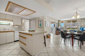 A modern kitchen with a central island, a dining area with a glass table, and a view of a residential building. - South Maui Beach Getaway | 2BR Condo Across from Kamaole Beach II (Kihei)
