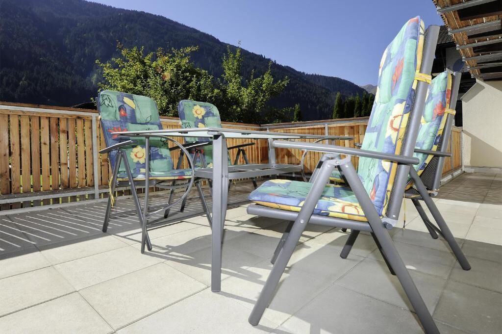 Eine Terrasss mit Tisch und zwei Stühlen, ein Holzzaun und eine Berglandschaft im Hintergrund.