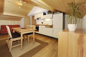 Una cocina compacta con gabinetes blancos, microondas y lavamanos. Hay una zona de comedor con una mesa y sillas. Una planta en maceta está sobre una encimera de madera.