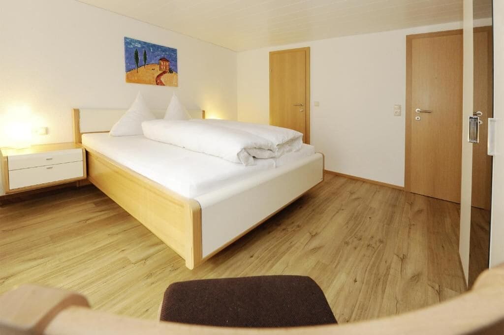 A bedroom with a bed, a nightstand, a wooden floor, and a door.