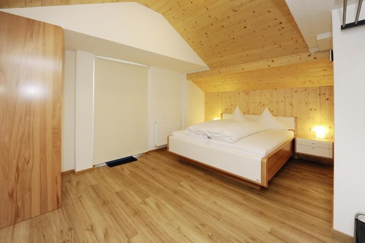 Un dormitorio con techo de madera, una cama con ropa blanca y una mesita de noche con lámpara.