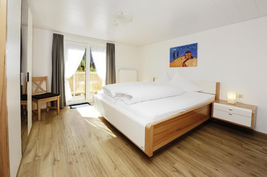A bedroom with a wooden bed, a bedside table, a chair, and a window with curtains.