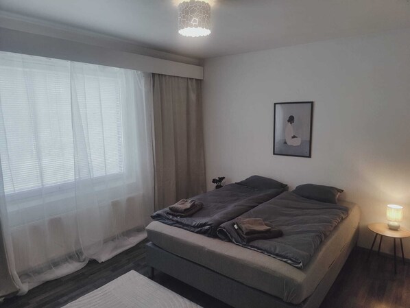 A bedroom with a bed, a bedside lamp, a framed picture on the wall, and a window with curtains.
