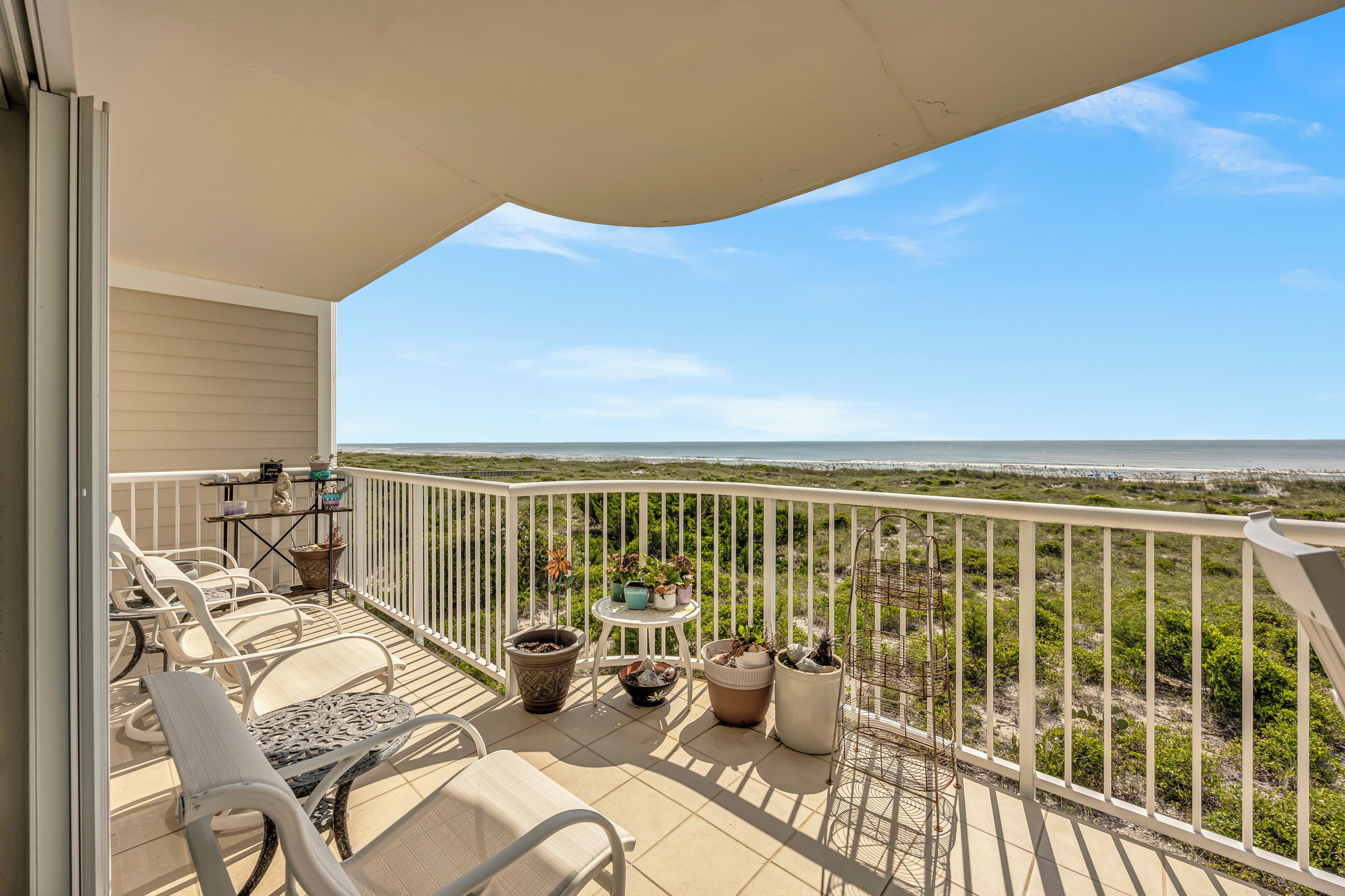 A balcony with a view of the beach, white outdoor furniture, and potted plants.