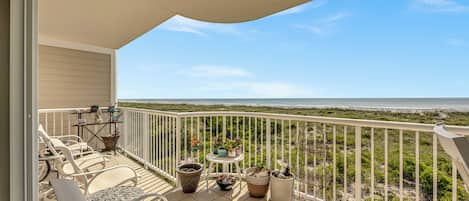A balcony with a view of the beach, white outdoor furniture, and potted plants.