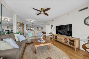 A spacious living room with a ceiling fan, a large flat-screen TV, a wooden coffee table, and a beige sofa. - Updated oceanfront 3br/3bath condo at Wrightsville Dunes! (Wrightsville Beach)