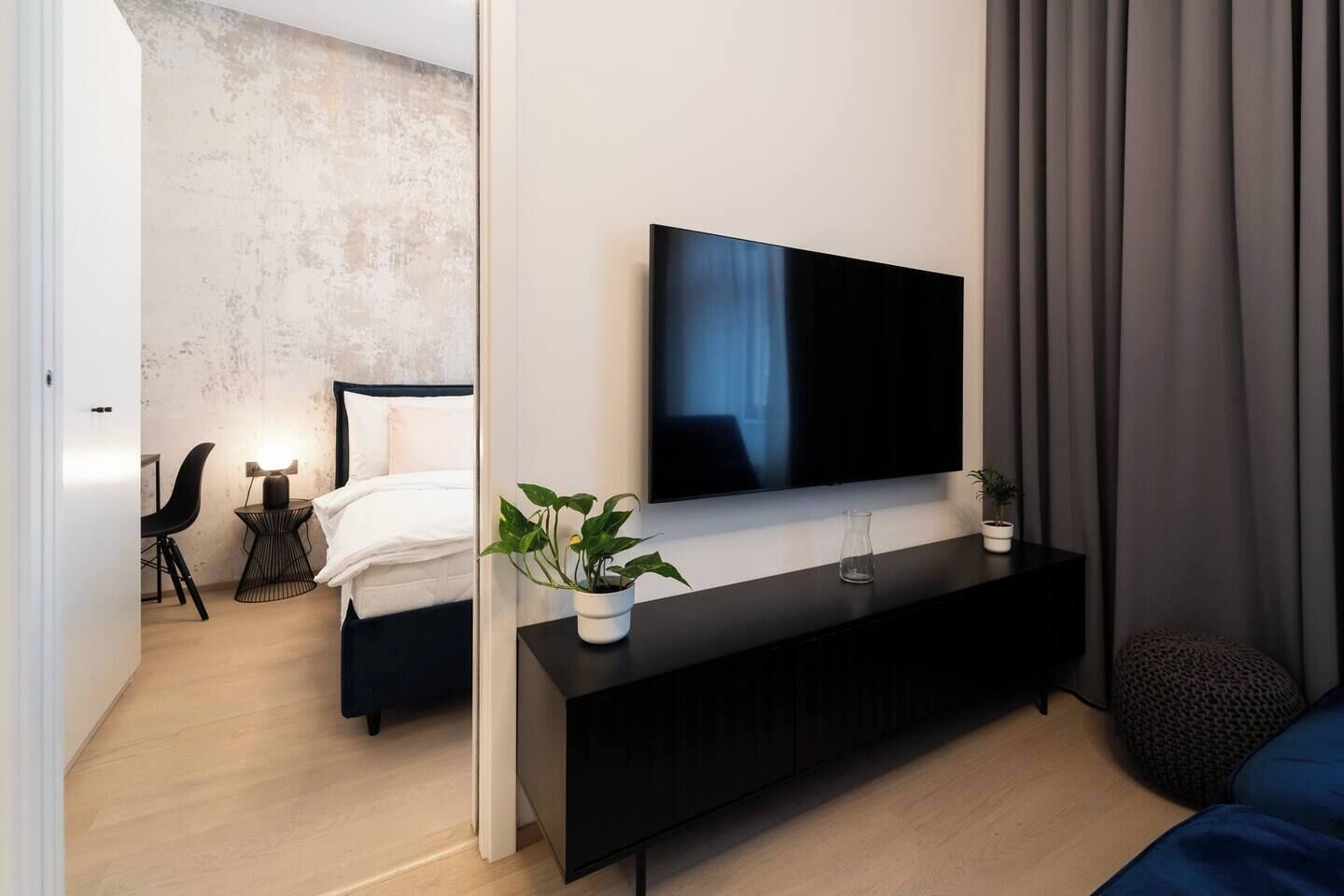 Ein modernes Hotelzimmer mit einem großen Flachbildfernseher an einer schwarzen Konsole, einem Bett mit weißer Bettwäsche, einem kleinen Nachttisch mit einer Lampe und einer Zimmerpflanze.