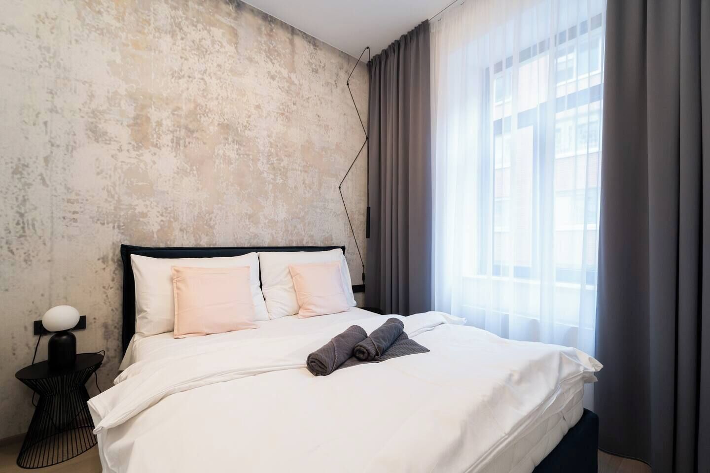 Hotellihuone, jossa on sänky, yöpöytä lampun kanssa ja ikkuna, jossa on verhot.