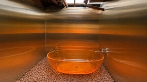 Una habitación con una bañera de vidrio grande y transparente llena de agua, rodeada de un piso de guijarros y vistas al cielo a través de una ventana.