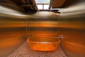 Ein Zimmer mit einer großen, transparenten Glasbadewanne, gefüllt mit Wasser, einem Kieselboden und Blick zum Himmel durch ein Fenster.