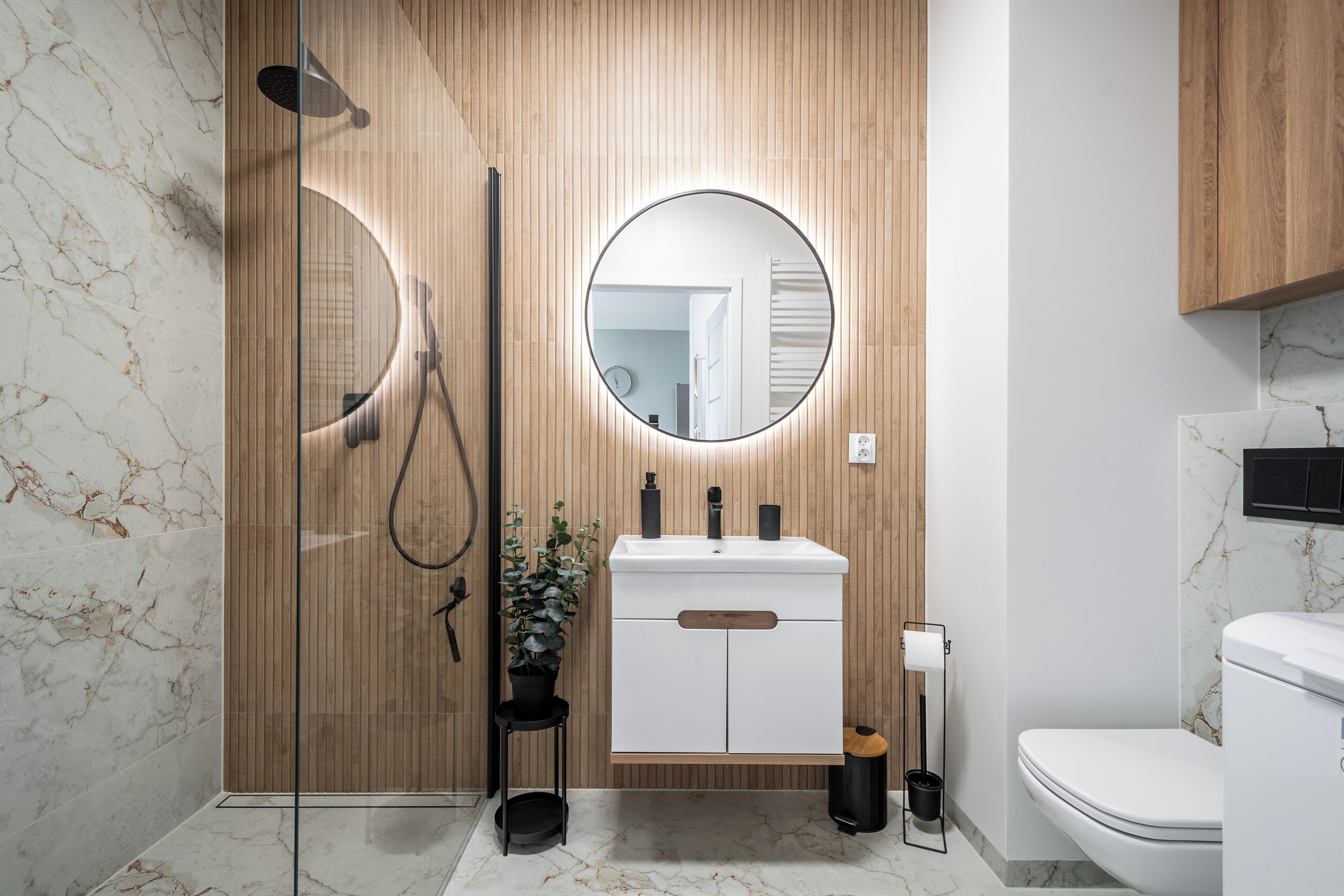 A modern bathroom with a wooden vanity, a round mirror, a shower with a handheld showerhead, and a toilet.