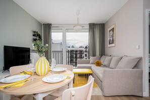 Una sala de estar moderna con sofá, una mesa de comedor dispuesta con platos y vasos, televisor y una ventana con cortinas.