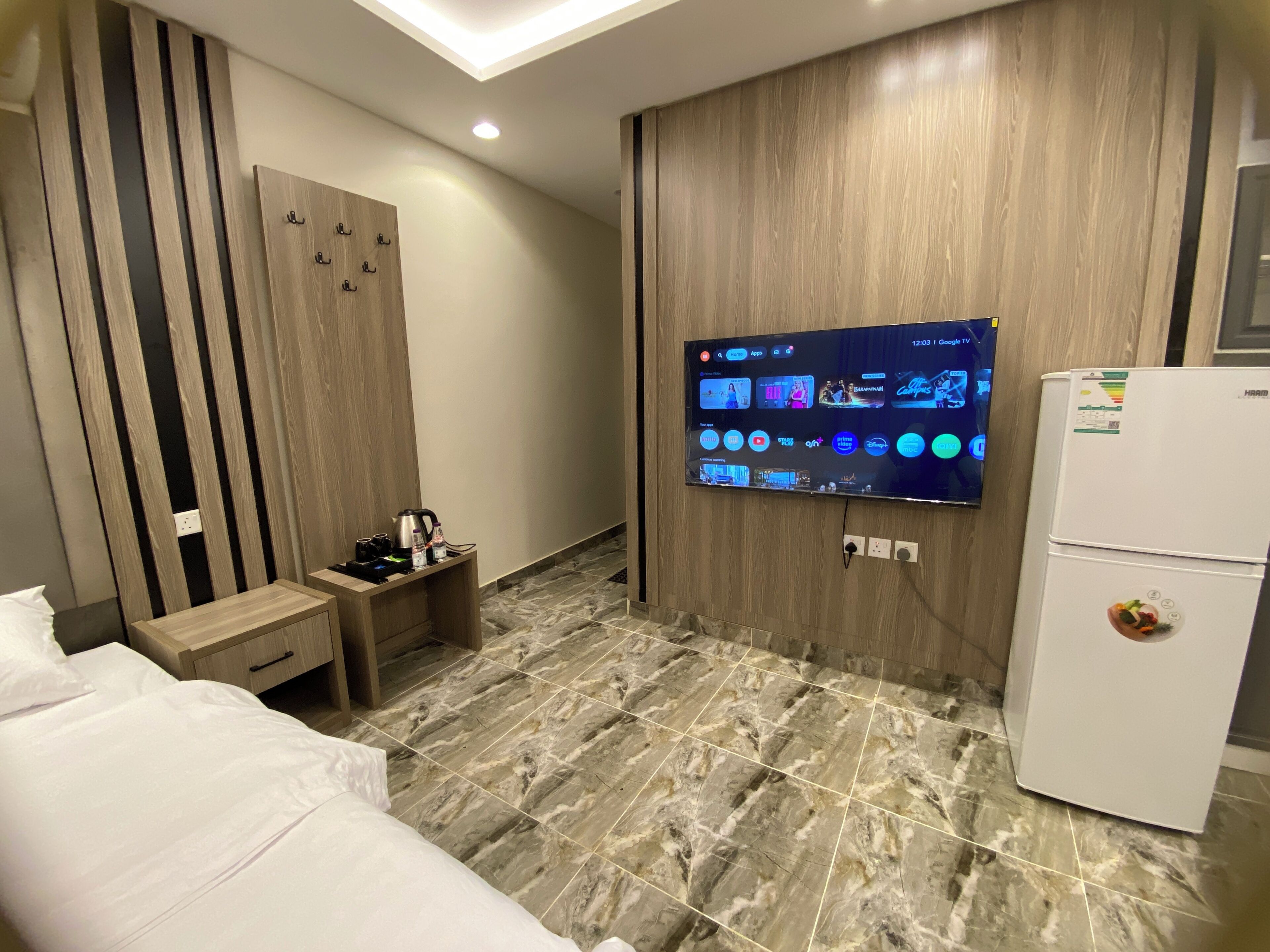 A hotel room with a bed, a TV mounted on the wall displaying various channels, a small wooden table with a kettle, and a refrigerator.