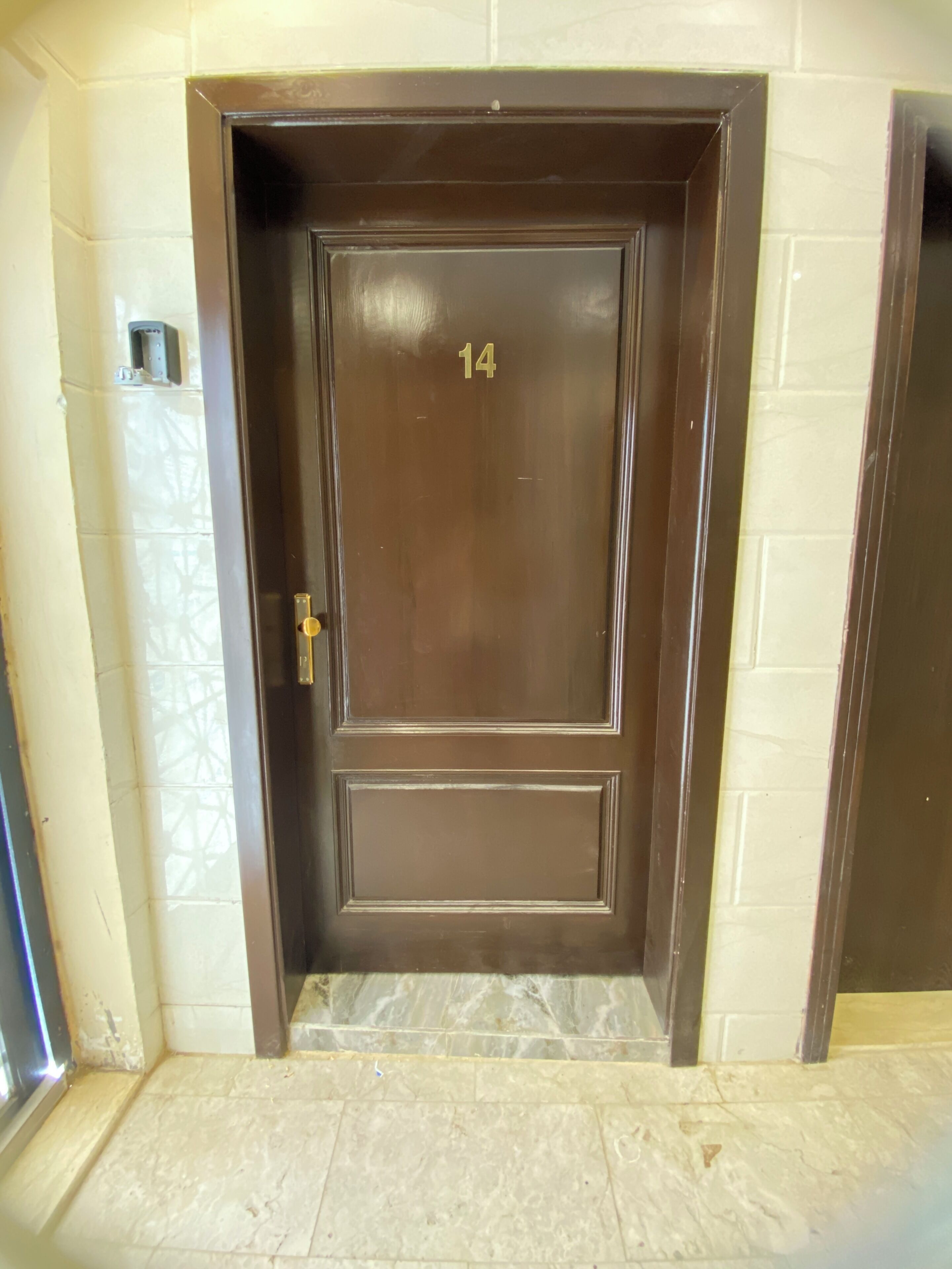 Una puerta marrón con el número 14, un escalón que lleva a ella y un dispositivo fijado a la pared a la izquierda.
