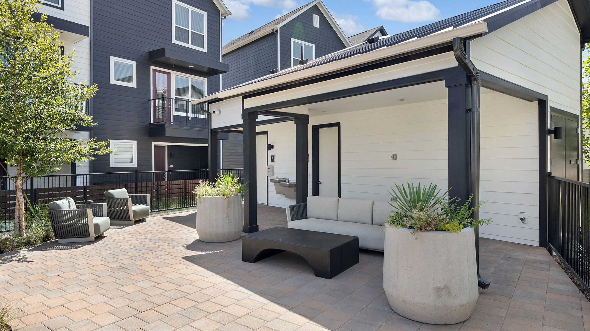 A covered outdoor seating area with a bench and planters, adjacent to a building with a dark exterior and white siding.