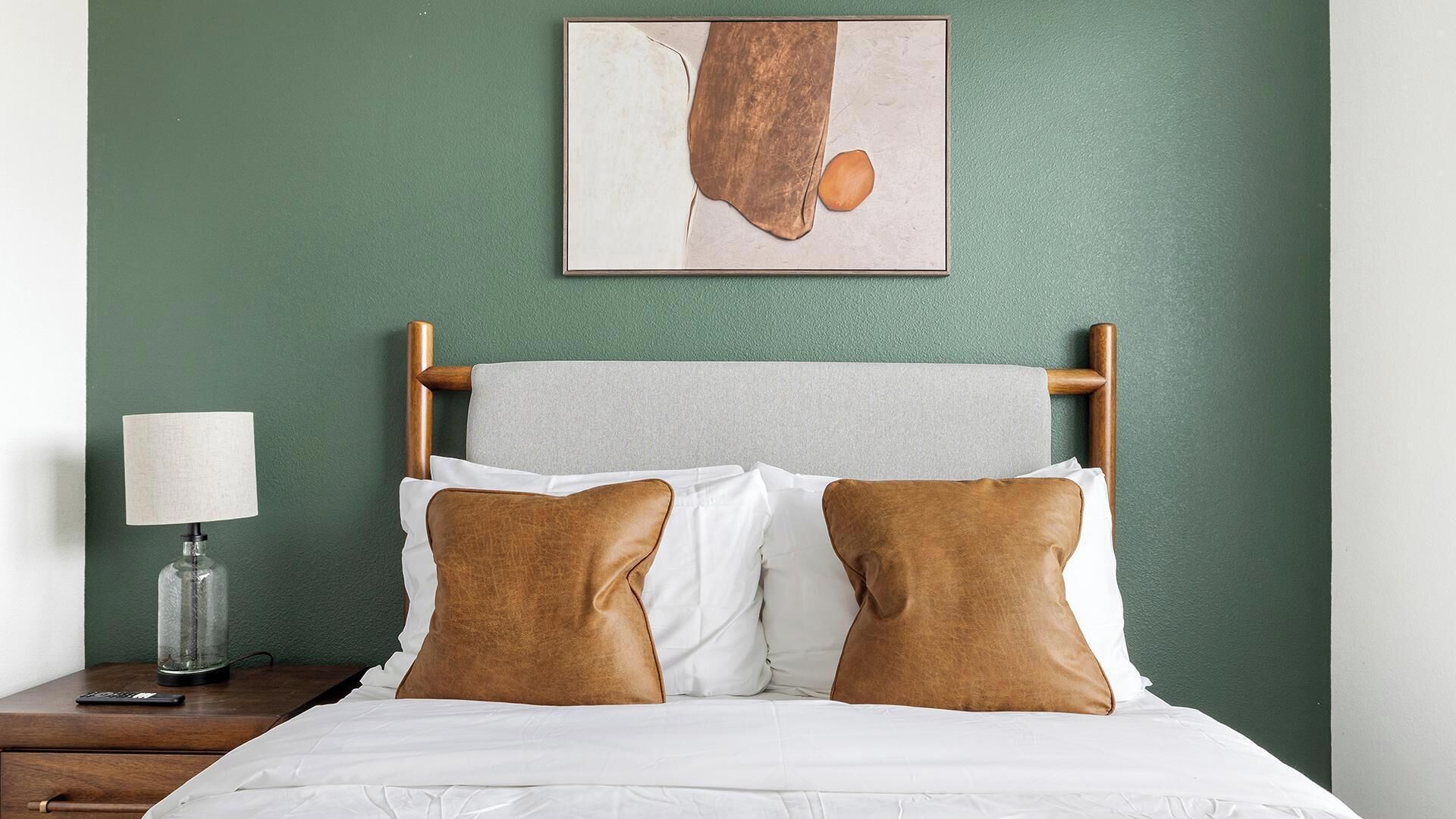 Een bed met witte lakens en twee bruine kussens, een nachtkastje met een lamp, en een ingelijst foto aan de muur.