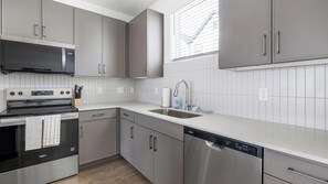 A modern kitchen with grey cabinets, stainless steel appliances, and a window with blinds. - Landing Amazing 2bd, Courtyard, Pool (Dallas)