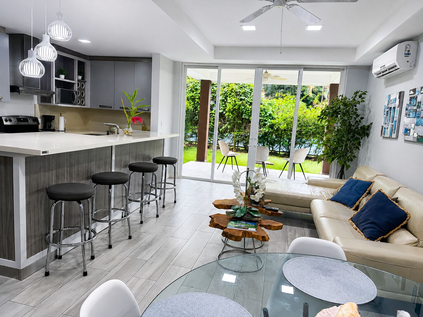 A modern kitchen with a bar area, a dining table with chairs, and a sofa in a living area with a view of greenery.