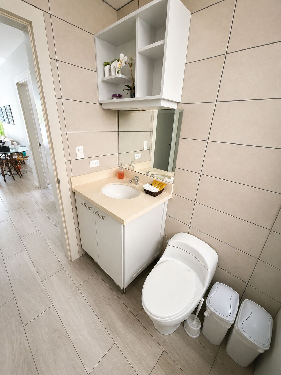 Une salle de bain moderne avec un lavabo blanc, un miroir et des toilettes. Une étagère se trouve au-dessus du lavabo, où sont posés des articles de toilette. Le sol est carrelé et une poubelle est placée à côté des toilettes.