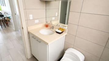 Une salle de bain moderne avec un lavabo blanc, un miroir et des toilettes. Une étagère se trouve au-dessus du lavabo, où sont posés des articles de toilette. Le sol est carrelé et une poubelle est placée à côté des toilettes.