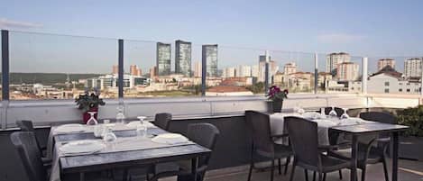 An outdoor dining area with tables and chairs, a city view, and a potted plant.