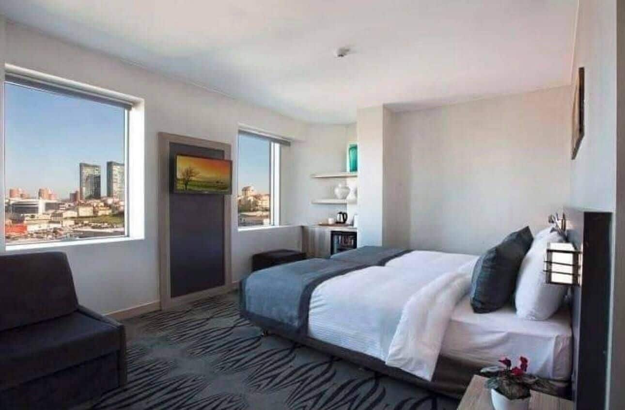 Quarto de hotel com cama, escrivaninha, cadeira, janela e vista para a cidade, e porta.