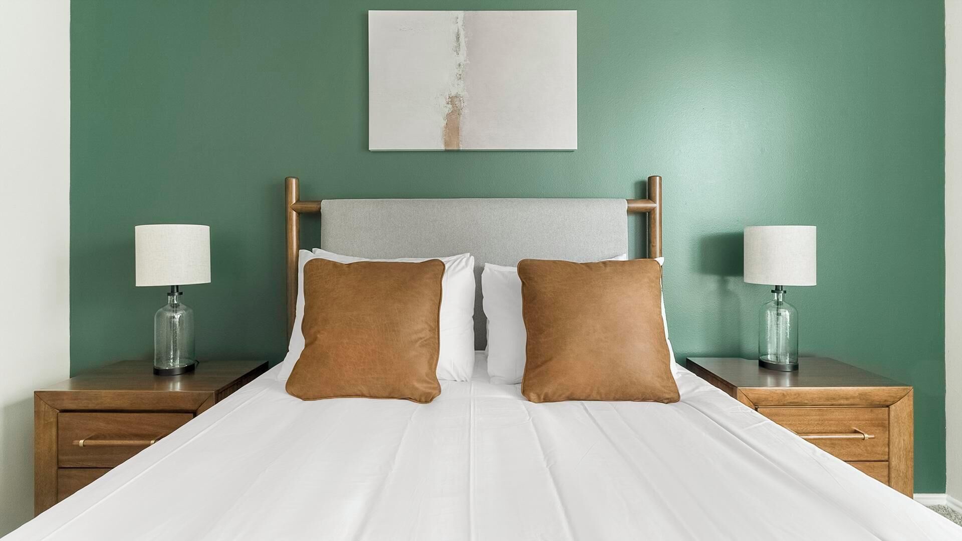 A bed with white linens, two brown pillows, and a wooden headboard. There are two bedside tables with lamps and a wall-mounted artwork above the bed.