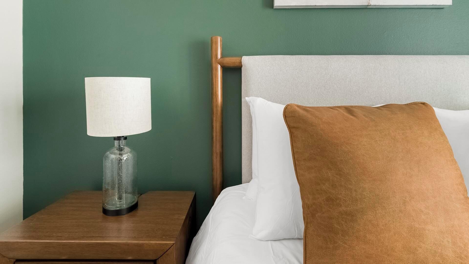 Ein ordentlich bezogenes Bett mit weißen Leinen und einem braunen Kopfkissen, eine klare Glaslampe auf einem hölzernen Nachttisch und eine grüne Wand.