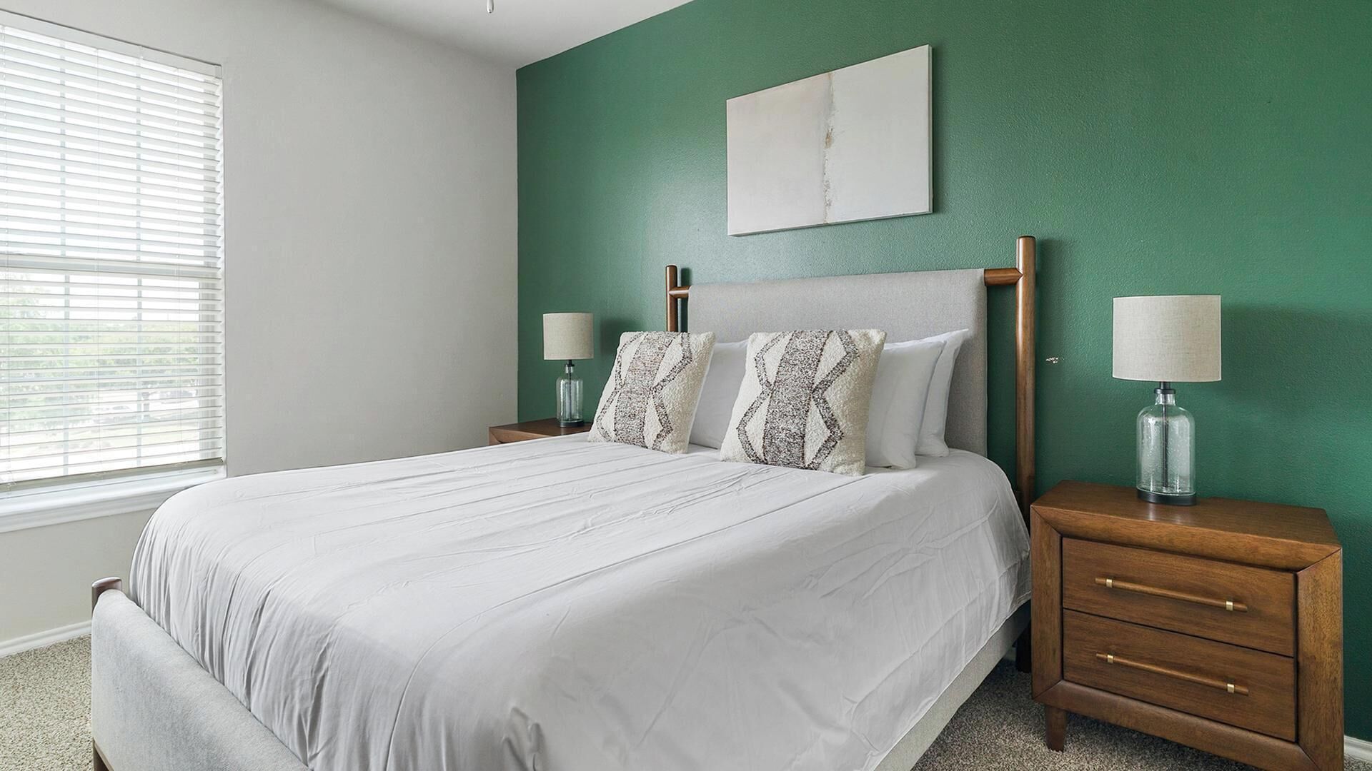 Une chambre à coucher avec un lit, deux tables de chevet, une lampe, une fenêtre avec des stores et un mur vert.