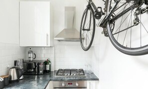 A kitchen with white cabinets, a gas stove, a coffee maker, and a bicycle hanging from the ceiling. - Light filled studio with view of Regents Park. 1 min walk to Baker St tube. (London)