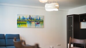 Ein Wohnzimmer mit einem Gemälde von einem See und Bäumen, einem Sofa, einem Esstisch mit Stühlen und einem Schrank.