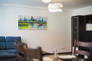 A living room with a painting of a lake and trees, a sofa, a dining table with chairs, and a cabinet. - Homely Apartment with Balcony - Central BA (Bratislava)