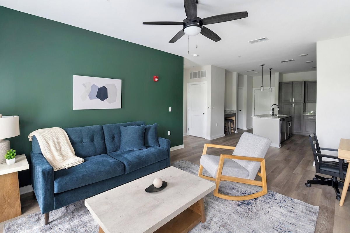 A modern living room with a blue sofa, a wooden coffee table, a chair, a ceiling fan, and a kitchen in the background.