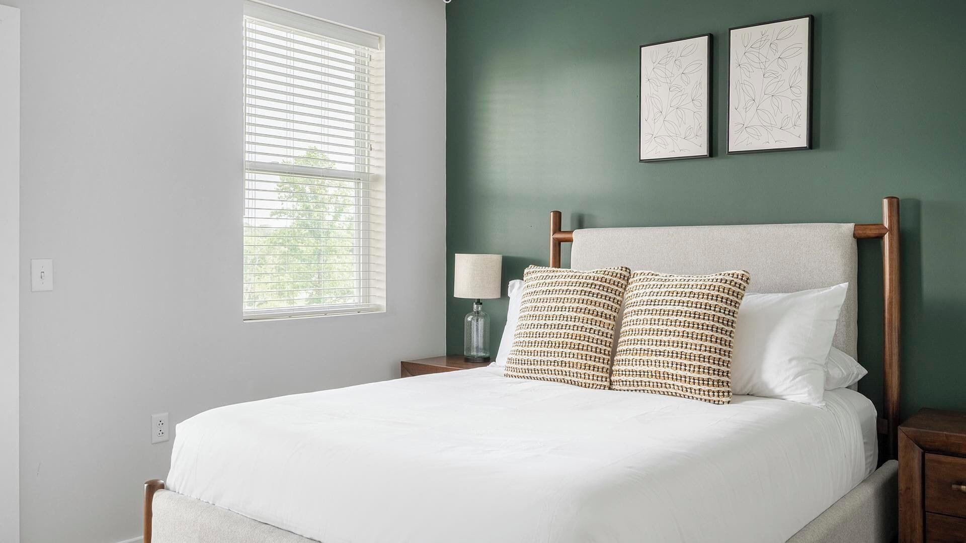 A bedroom with a bed, two wall-mounted picture frames, a nightstand with a lamp, and a window with blinds.