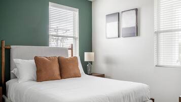 A bedroom with a bed, two windows with blinds, a nightstand with a lamp, and two wall-mounted artworks.