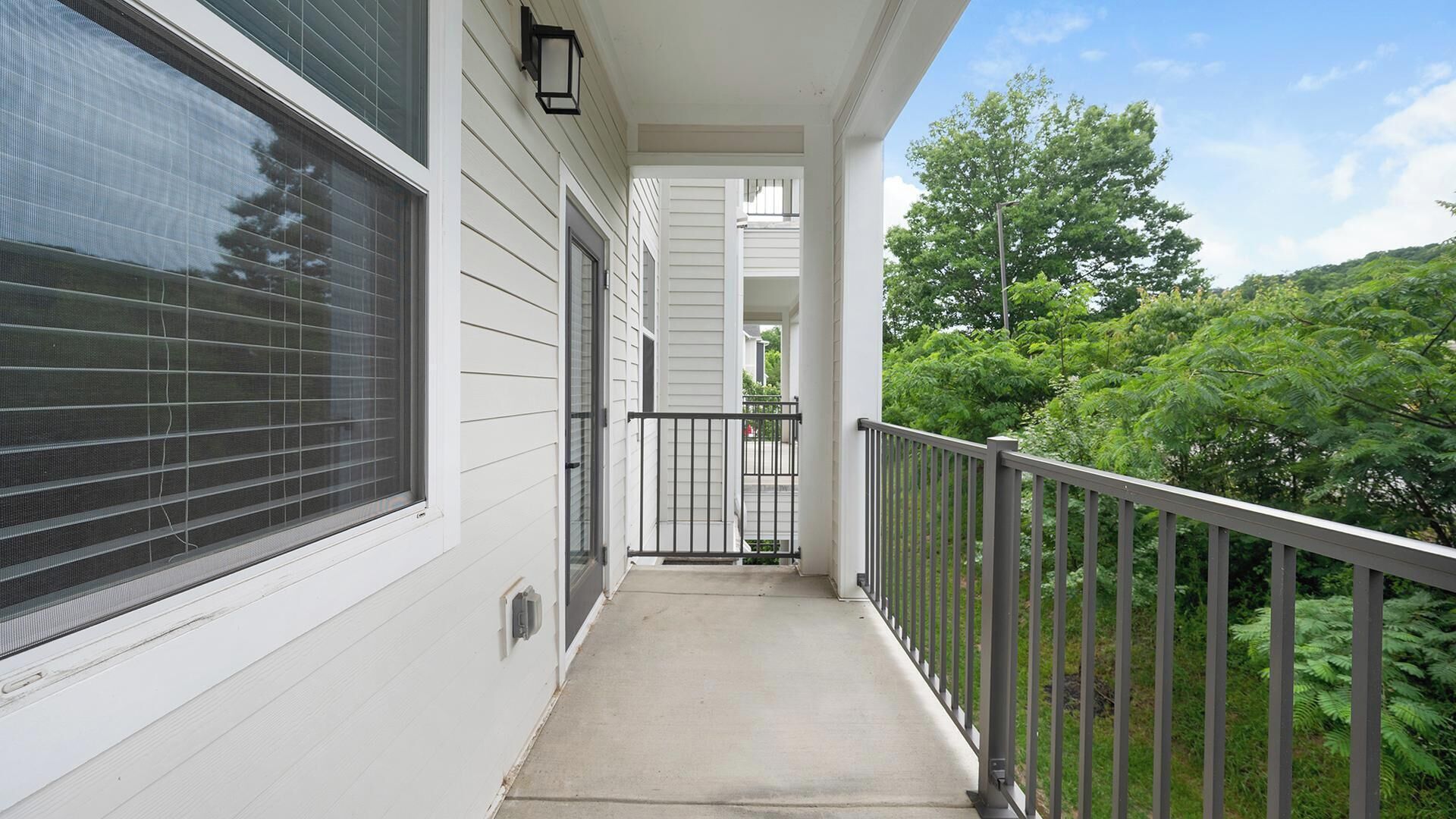 A covered balcony with a metal railing, a window with blinds, and a view of a green landscape.