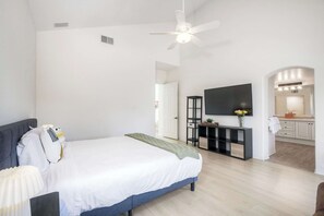Ein modernes Schlafzimmer mit einem großen Bett, einem Deckenventilator, einem Fernseher und einem durch einen Bogen sichtbaren Badezimmer.
