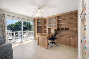 A spacious room with a large desk, ample shelving, a ceiling fan, and a balcony with outdoor furniture. - Brick & Beach Family Retreat (Carlsbad)